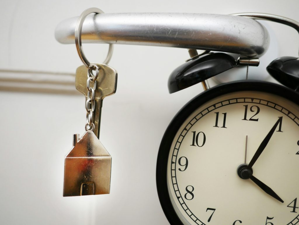 House key with small home keychain and clock hanging from a doorknob, representing flexible closing timeline