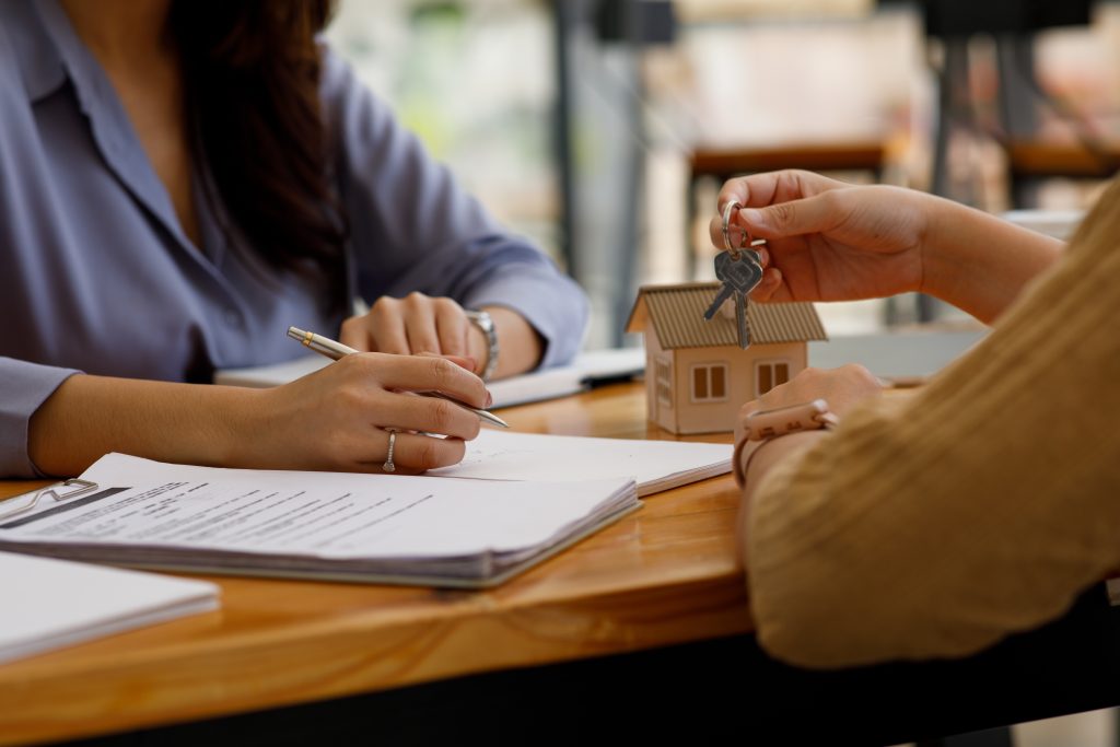 Hands exchanging house keys over a real estate closing table with paperwork and a model home.