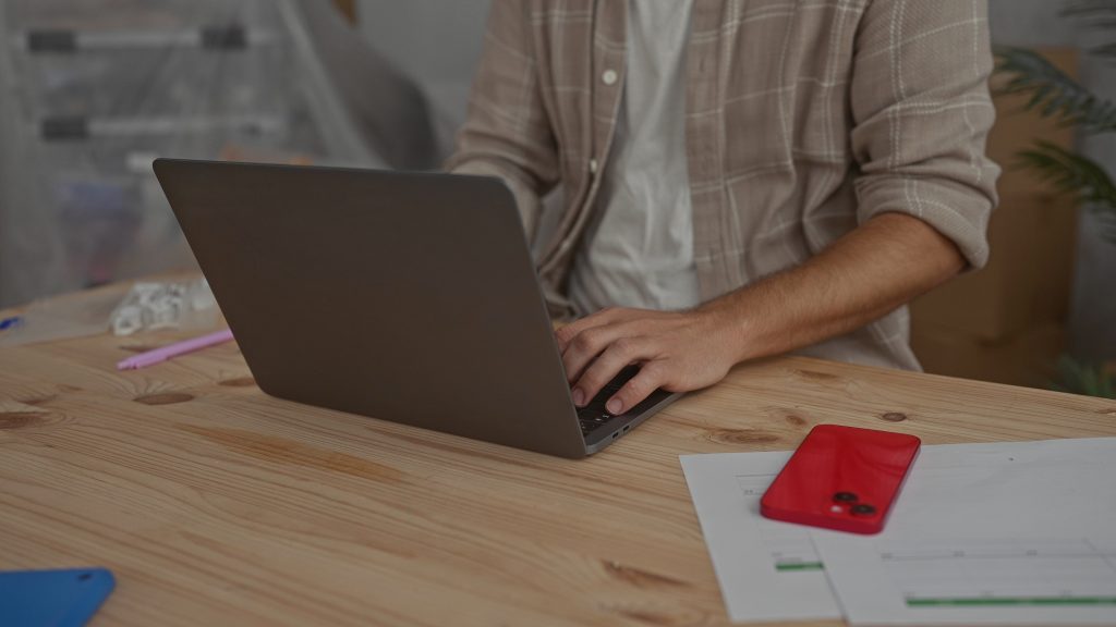 Homeowner using a laptop at home with paperwork and a phone nearby while reviewing home selling options.
